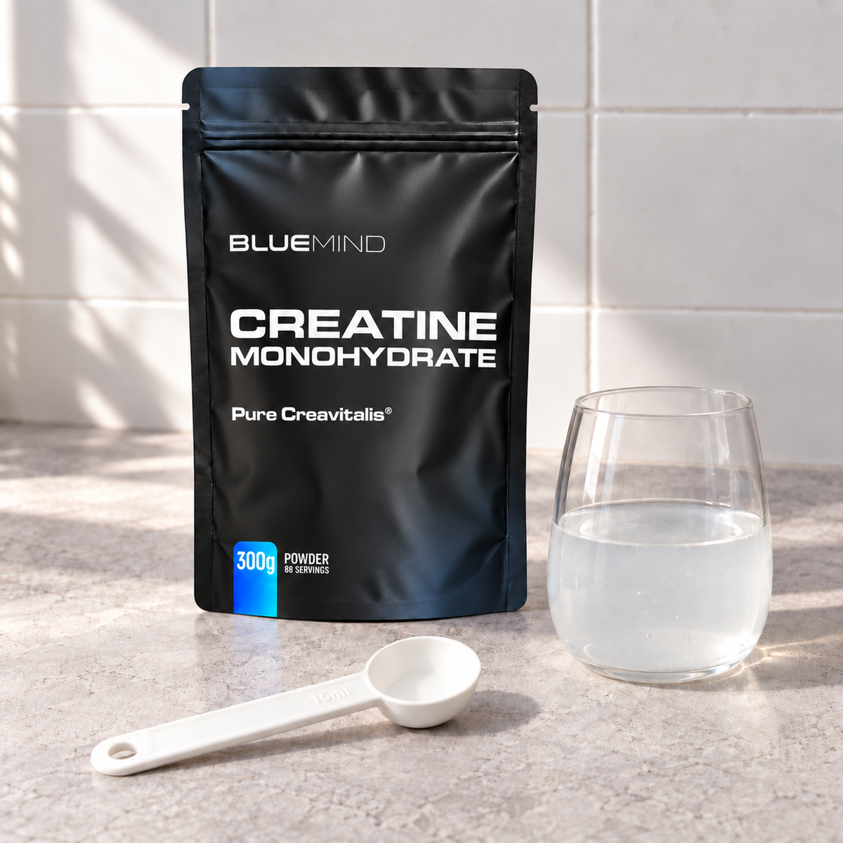 BLUEMIND Kreatin Monohydrat Creavitalis® – 300-g-Beutel mit Messlöffel und Glas Wasser auf Arbeitsplatte